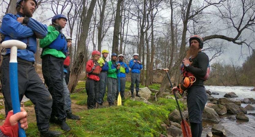 A group of people wearing lifejackets and helmets stand on the bank of a river, listening to an instructor. 